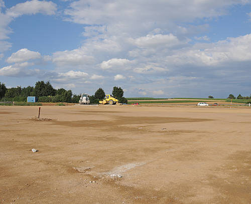The site is cleared, flattened by bulldozers and ready for development. Building area in the country. Farmland had to yield, the soil will be sealed. The ground has been levelled and prepared for rising a factory or new homes. Wide, empty space. Blue cloudy sky. Trees, bushes, highway and construction machinery in the distance. Horizontal orientation.